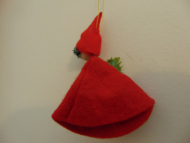 May include: A red felt Christmas ornament shaped like a person wearing a red pointed hat and a green holly sprig.