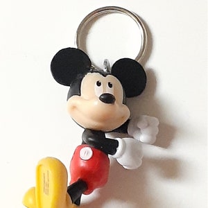 May include: A black and yellow Mickey Mouse keychain with a red shirt and white gloves. The keychain has a silver metal ring.