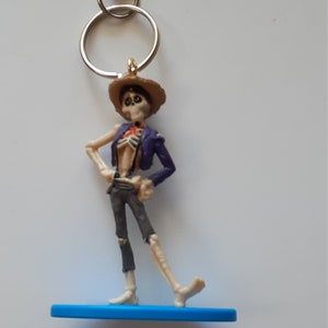 Könnte beinhalten: Ein blauer und weißer Skelett-Figuren-Schlüsselanhänger mit einem braunen Hut und einer lila Jacke. Die Figur steht auf einem blauen Sockel mit einem silbernen Schlüsselring.