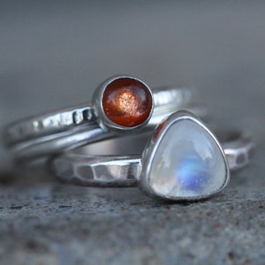 May include: Three silver rings with gemstone settings. One ring features a round, orange-brown stone, while another has a triangular, iridescent white stone. The rings have textured bands.
