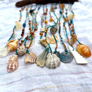 May include: A wall hanging featuring multiple strands of teal and brown cord suspended from a piece of driftwood. The strands are decorated with various seashells, small glass beads in amber and blue tones, and a circular copper-toned metal disc.