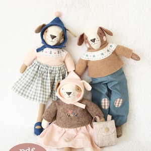 Könnte beinhalten: Drei Plüsch-Hunde-Spielzeuge mit gestickten Details. Der Hund links trägt einen blauen Hut, einen rosa Pullover, einen karierten Rock und blaue Schuhe. Der Hund in der Mitte trägt einen braunen Pullover, einen rosa Rock und blaue Schuhe. Der Hund rechts trägt einen braunen Pullover, eine blaue Hose und blaue Schuhe. Die Hunde sitzen alle auf einer weißen Oberfläche. Der Text "pdf pattern" ist in der linken unteren Ecke des Bildes sichtbar.