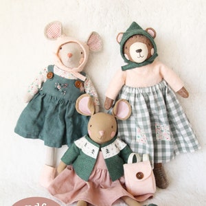 May include: Three handmade plush toys: a brown bear wearing a green hat and a pink dress with a gingham skirt, a mouse wearing a pink hat and a green dress, and a mouse wearing a green jacket and a pink dress with a pink backpack.