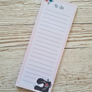 May include: A light pink to-do list notepad with a floral design and a black sewing machine illustration. The notepad has lined pages and the words "to do" at the top. The notepad is rectangular and sits on a wooden surface.