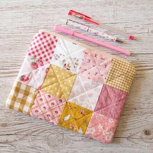 May include: A patchwork fabric pencil case with a variety of pink, yellow, and white patterns. The case is made of quilted fabric and has a zipper closure.