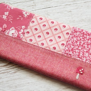 May include: A pink patchwork pencil case with a zip closure. The case features floral, mushroom, and patterned fabric panels. A small embroidered mushroom embellishes the bottom corner. The case is made of various pink fabrics.
