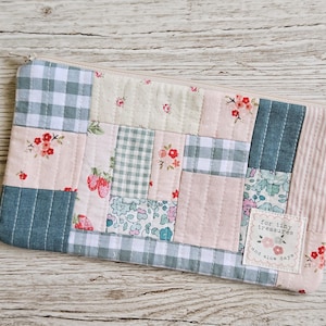 May include: A rectangular zippered pouch made of quilted fabric featuring a patchwork design of various patterns including green and white gingham, floral prints, and strawberry motifs. A small white label reads for tiny treasures and slow days.
