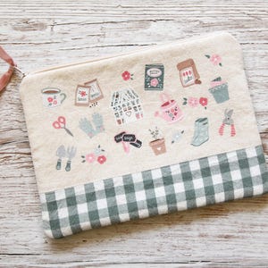 May include: A beige zippered pouch with gardening-themed illustrations. The pouch features a green and white checkered bottom panel and a pink ribbon pull. The illustrations include gardening tools, seeds, and flowers.
