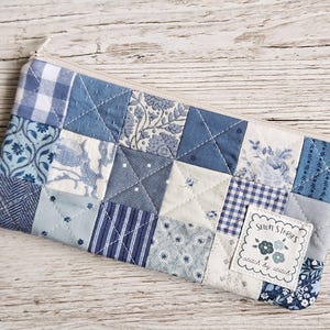 May include: A rectangular zippered pouch made of quilted fabric featuring a patchwork design of various blue and white patterns including gingham, floral, and stripes. A white fabric label is sewn on the front with the text Sewn Stories stitch by stitch.