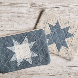 May include: Two rectangular zippered pouches featuring quilted star patterns. One pouch is dark blue with a light floral star center, while the other is light beige with a dark blue star. Both items display intricate geometric stitching throughout.
