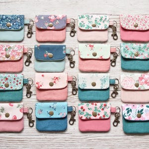 Peut inclure: Collection de petites pochettes en tissu colorées avec des motifs floraux et des porte-clés. Chaque pochette présente une combinaison de couleurs différente, dont le rose, le bleu et le vert, avec une fermeture à bouton. Les pochettes sont disposées sur une surface en bois clair.