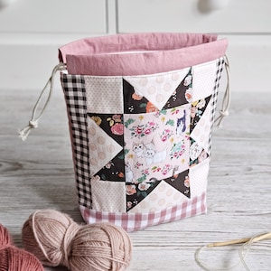 Könnte beinhalten: Ein Patchwork-Garnbeutel mit rosa Innenfutter und Kordelzug. Die Außenseite zeigt ein Sterndesign mit Blumen- und Katzenstoff, ergänzt durch schwarz-weißes Vichy- und Tupfenmuster. Zwei Wollknäuel und Stricknadeln sind sichtbar.