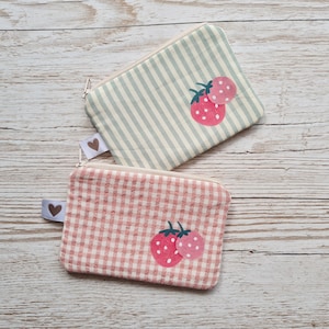 Könnte beinhalten: Zwei kleine Reißverschlusstaschen mit Erdbeermotiven. Eine Tasche hat ein hellgrünes und weißes Streifenmuster, die andere ein rosa-weißes Vichy-Muster. Jede Tasche zeigt eine rosa Erdbeere mit weißen Punkten.