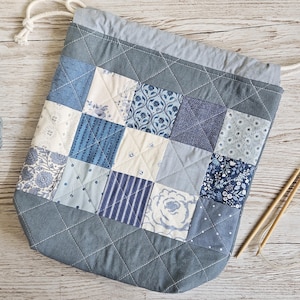May include: A quilted drawstring bag featuring a patchwork design of various blue and white fabric squares. The bag has a solid grey border, a grey top casing, and white rope drawstrings. The quilted pattern consists of diagonal stitching.