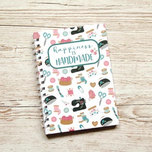 Könnte beinhalten: Ein spiralgebundenes Notizbuch mit weißem Einband, das ein sich wiederholendes Muster von Nähillustrationen in Rosa, Türkis und Schwarz aufweist. Der Einband enthält den Text "happiness is handmade" in einem türkis-weißen Design.
