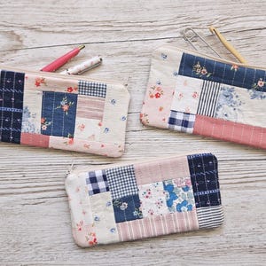 Könnte beinhalten: Drei handgefertigte Patchwork-Federmäppchen mit Reißverschluss. Jedes Etui hat eine einzigartige Kombination aus floralen, gestreiften und karierten Stoffflicken in Rosa, Blau und Weiß. Ein Stift und eine Häkelnadel sind sichtbar.