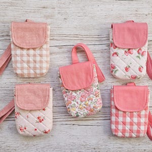 May include: A collection of pink and white quilted phone cases with matching wrist straps. The cases feature various patterns, including gingham, floral, and strawberry designs. Each case has a flap closure.