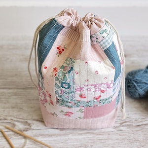 Könnte beinhalten: Ein gesteppter Beutel mit Kordelzug im Patchwork-Design aus verschiedenen Blumen- und Erdbeermustern in Rosa-, Blau- und Weißtönen. Der Beutel hat einen einfarbigen hellrosa Boden und einen passenden Kordelzugverschluss mit zwei beigen Kordeln.