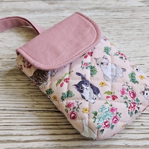 May include: A quilted pink eyeglasses case with a matching strap and flap closure. The case is decorated with a repeating pattern of kittens, flowers, and foliage in shades of pink, white, and green. The case is on a wooden surface.