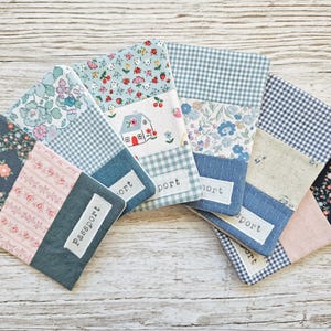 Könnte beinhalten: Eine Auswahl an Patchwork-Passhüllen mit verschiedenen Blumen- und Vichy-Mustern. Die Hüllen haben eine Mischung aus blauen, rosa und marinefarbenen Stoffen. Einige haben ein weißes Etikett mit dem Aufdruck "Passport".