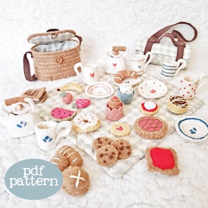 May include: A selection of felt food items, including a picnic basket, mugs, plates, and pastries, arranged on a checkered cloth. The text "pdf pattern" is visible. The items include a croissant and a strawberry.