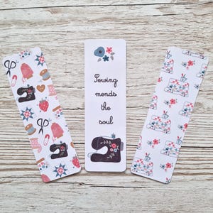 May include: Three white bookmarks with a sewing theme. One bookmark features a sewing machine and flowers, another has a pattern of fabric scraps and flowers, and the third has the text "Sewing mends the soul".