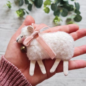 Könnte beinhalten: Ein weißes Filz-Schaf mit einem rosa Band und einer kleinen Glocke. Das Schaf liegt auf der Seite mit ausgestreckten Beinen.
