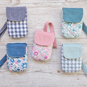 May include: Five quilted fabric phone cases in various patterns and colors. The cases feature a flap closure and a wrist strap. Patterns include gingham, floral, and solid colors like blue and pink. The cases are arranged on a light wood surface.
