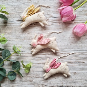 May include: Three handmade fabric bunny ornaments with a white background and a gingham bow. The bows are yellow, pink, and red.