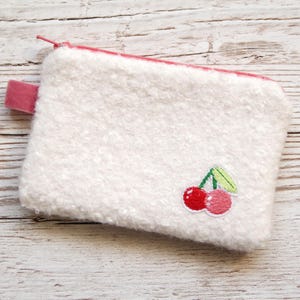 Könnte beinhalten: Eine weiße, strukturierte Tasche mit einem rosa Reißverschluss und einer kleinen rosa Stoffschlaufe. Die Tasche hat ein gesticktes Design von zwei roten Kirschen mit grünen Blättern. Die Tasche ist rechteckig.