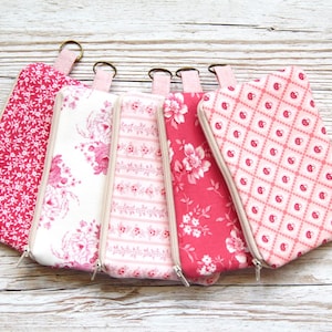 May include: Five fabric pencil cases in pink and white, each with a different floral or patterned design. The cases have a zip closure and a small loop at the top. The cases are arranged on a light wood surface.