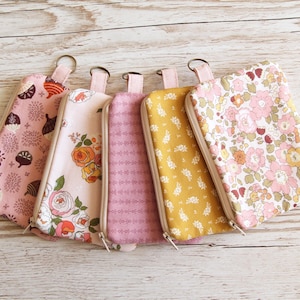 May include: Five fabric zippered pouches in various floral and patterned designs. The pouches are in shades of pink, yellow, and purple, with a small loop at the top. The background is a light wood surface.
