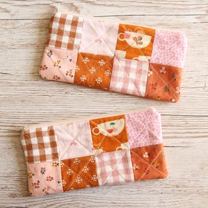 May include: Two quilted pencil cases with a patchwork design. The cases feature squares of pink, orange, and brown fabrics with floral, gingham, and teacup patterns. The cases are rectangular with a zip closure.