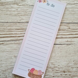 May include: A rectangular notepad with a pink border and lined paper. The top features the words "to do" and floral accents. The bottom has a basket illustration filled with yarn balls in pink, peach, and cream colours.