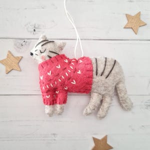 May include: A grey and white felt cat wearing a red jumper with white hearts and a white string for hanging.
