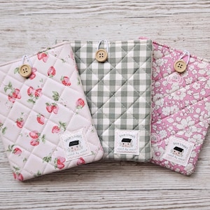 May include: Three quilted fabric cases in pastel shades. One has a strawberry pattern, another a grey and white gingham check, and the third a pink floral design. Each case has a wooden button closure and a label reading "Sewn Stories".
