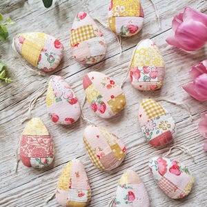 May include: Twelve handmade patchwork fabric Easter eggs in pink, yellow and white with floral and gingham patterns. The eggs are strung together with white twine.