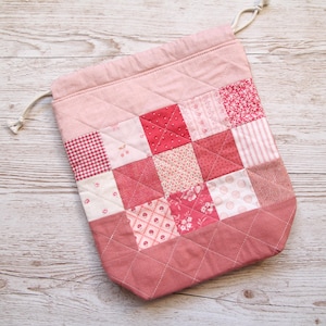 May include: A pink quilted drawstring bag with a patchwork design. The patchwork features squares of various pink and red floral and patterned fabrics. The bag has a diamond-stitched bottom and a drawstring closure with a beige cord.