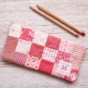 May include: A pink and white patchwork pencil case with a variety of fabric designs, including floral, gingham, and striped patterns. Two pencils are placed on the wooden surface next to the case. The case has a label that reads "Stitch Stories".