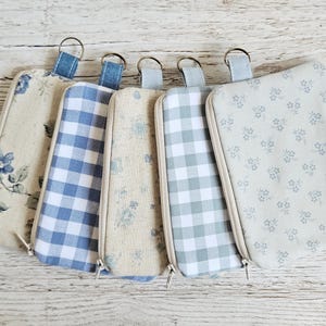 May include: Five rectangular fabric pouches with zippers and metal key rings. The pouches feature various patterns including blue floral prints on beige, blue and white gingham checks, and light blue floral designs on a light gray fabric background.
