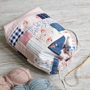 Peut inclure: Un sac à cordon patchwork en tissus roses, bleus et blancs. Le sac présente divers motifs, dont des motifs floraux, vichy et rayés. Deux pelotes de laine et des aiguilles à tricoter sont à côté du sac.