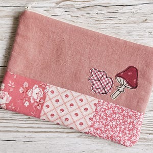 May include: A rectangular zipped pouch in dusty rose fabric featuring a patchwork base with floral, gingham mushroom, and small white floral patterns. An embroidered oak leaf and a red mushroom are appliqued on the upper section.