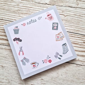 May include: A white notepad with a grey border and the word "notes" in black lettering. The notepad is decorated with a circle of gardening-themed illustrations, including a watering can, gardening gloves, a cup of tea, and a pair of gardening shears.