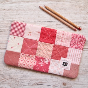 May include: A quilted pencil case in red and pink hues, with a patchwork design of different fabrics including gingham and floral prints. Two pencils are placed on a light wood surface. The case has a label that reads "Sew Very Happy".