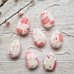 May include: Seven handmade fabric Easter eggs in pink, white and beige with floral and cherry patterns. The eggs are hanging from white string on a white wooden background.