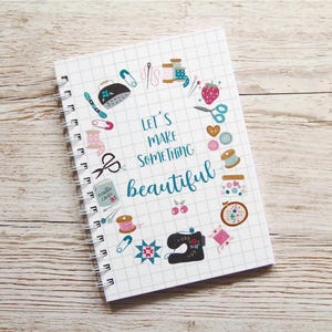 Könnte beinhalten: Ein weißes Spiralnotizbuch mit einem karierten Muster und dem Text "LET'S MAKE SOMETHING beautiful". Der Einband ist mit Illustrationen zum Thema Nähen verziert, darunter Scheren, Garnrollen und eine Nähmaschine.