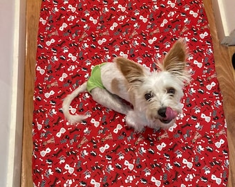 May include: A white dog wearing a green diaper is lying on a red dog bed with a pattern of black and white dogs and bones.