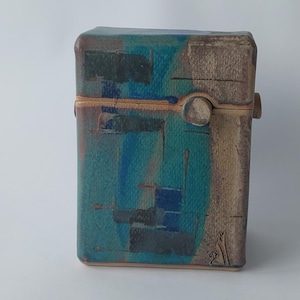 Op de afbeelding: Een keramische doos met een abstract ontwerp in blauw, groen en bruin. De doos heeft een scharnierend deksel en een kleine knop aan de voorkant.