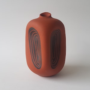 Puede incluir: Un jarrón de terracota con un diseño geométrico de círculos concéntricos negros. El jarrón tiene una forma rectangular con bordes redondeados.