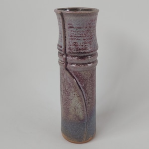 May include: A tall, cylindrical ceramic vase with a purple and gray glaze. The vase has a slightly flared top and a textured surface.